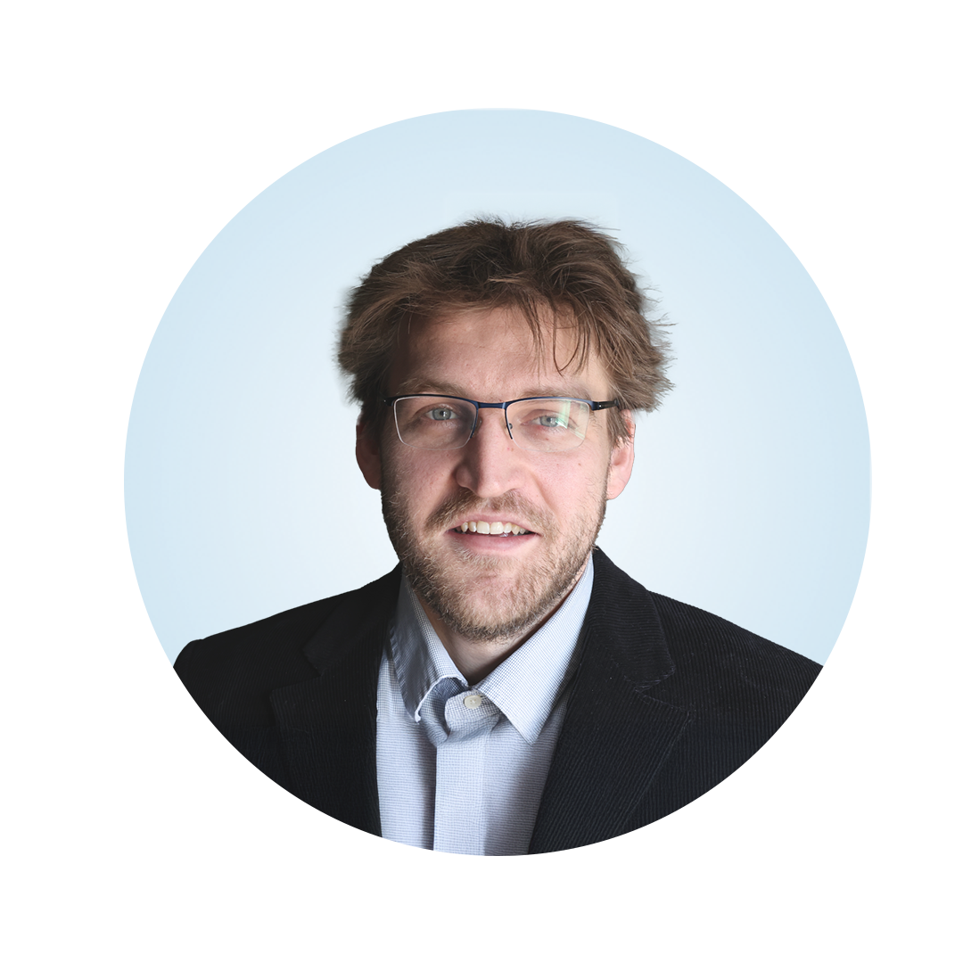 Head-and-shoulders portrait of a man with tousled light brown hair and glasses, wearing a dark blazer over a light collared shirt, against a light blue circular background.