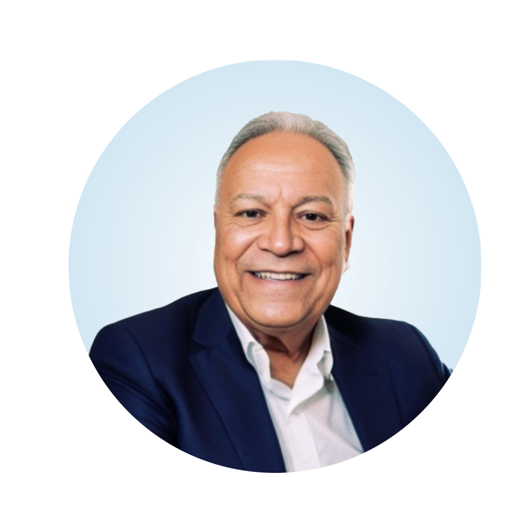 Head-and-shoulders portrait of a smiling older man with short gray hair, wearing a dark blazer over a white collared shirt, against a light blue circular background.