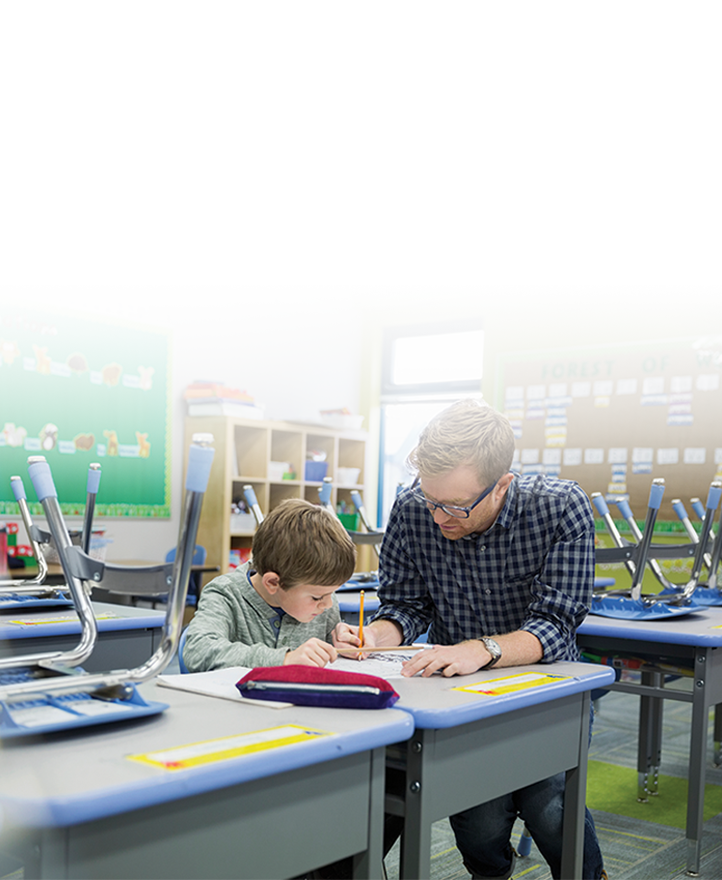 Teacher assisting a young student with classwork at a desk in an elementary classroom, providing one-on-one guidance.